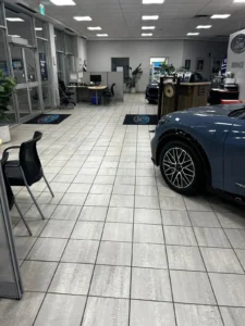 A car dealership showroom with a blue car parked inside. The space features tiled floors, glass doors, and several desks with chairs. Framed posters and signs with the Ford logo are visible on the walls.