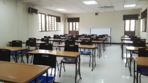 A classroom with multiple rows of empty desks and chairs. The room has white walls, large windows with blinds partially drawn, and a whiteboard at the front. The floor is shiny, and the lights are on, illuminating the space.