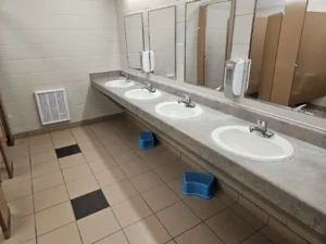 A clean public restroom with five white sinks and rectangular mirrors above each. Beneath two sinks are blue step stools. The floor has a tan tile design with black accents. Beige stalls are visible on the right.