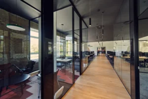 A modern office space featuring glass-walled meeting rooms on the left and an open workspace with desks and chairs on the right. Wooden flooring runs through the center, and large windows illuminate the area with natural light.