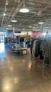 A spacious clothing store with bright lighting and polished concrete floors. Racks of hoodies and athletic wear are displayed. Several shoppers browse, and a colorful wall mural is visible in the background.