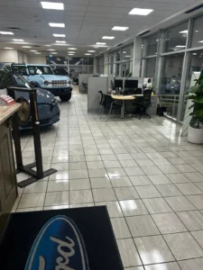 Car dealership showroom with cars on the left and office desks on the right. The floor is tiled, and there's a mat with the Ford logo in the foreground. Large windows line the right side, providing natural light.