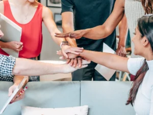 A diverse group of people pile their hands together in a show of teamwork while sitting on a sofa. They all have arms outstretched, joining hands in the center. The setting appears casual, with books and cushions around.
