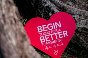 A large red heart-shaped sign leaning against a tree trunk with the motivational text "Begin today for a better tomorrow" and a heartbeat line graphic underneath. The scene conveys themes of health and inspiration.