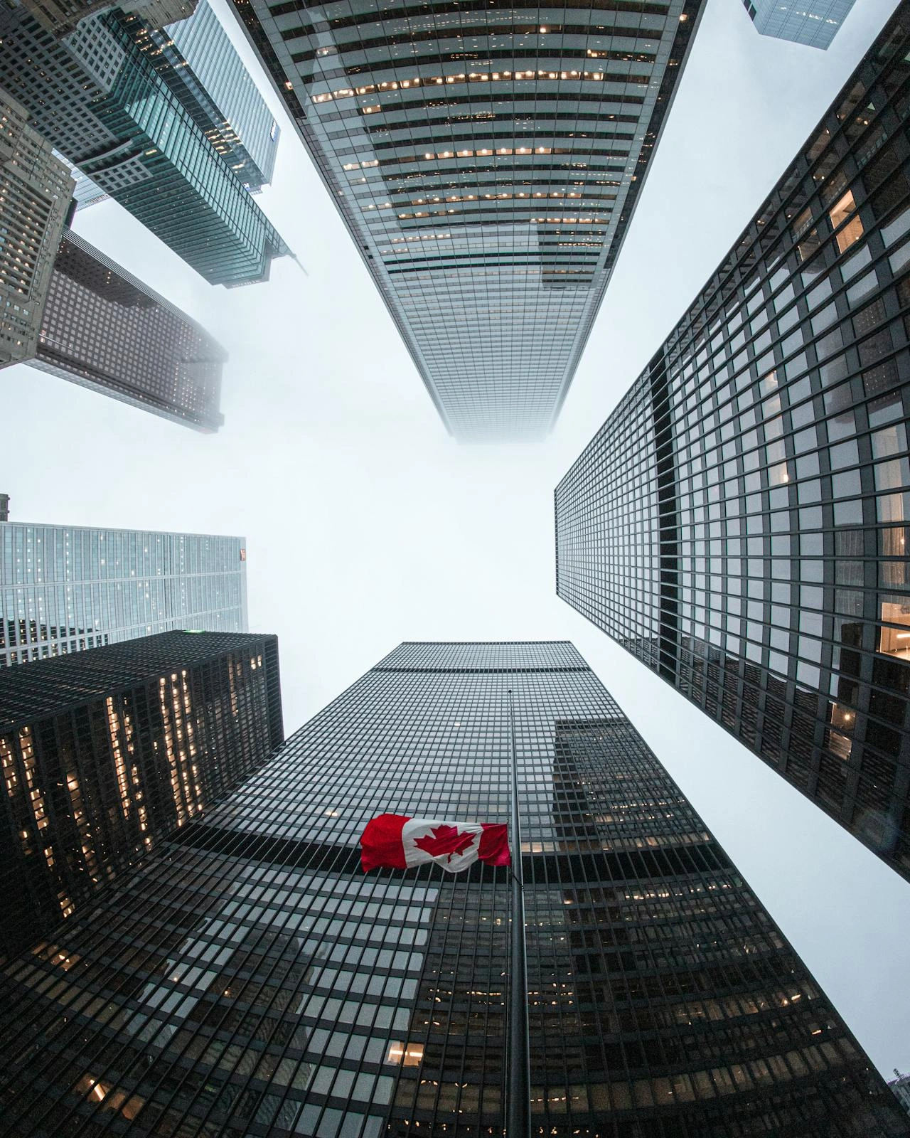 A low-angle view of tall skyscrapers in a city, with a Canadian flag on one of the buildings. The buildings create a symmetrical pattern, converging towards the cloudy sky.