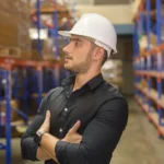 A man in a dark shirt and white hard hat stands with arms crossed in a warehouse. He gazes to the side, surrounded by shelves and stacked boxes.