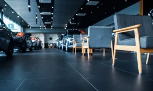 A modern car showroom interior featuring a row of sleek, dark upholstered chairs with wooden arms. Several cars are displayed in the background under bright spotlights on a polished dark floor.