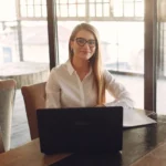 A person with long hair and glasses is smiling while sitting at a wooden table, using a laptop. They are in a well-lit room with large windows in the background.