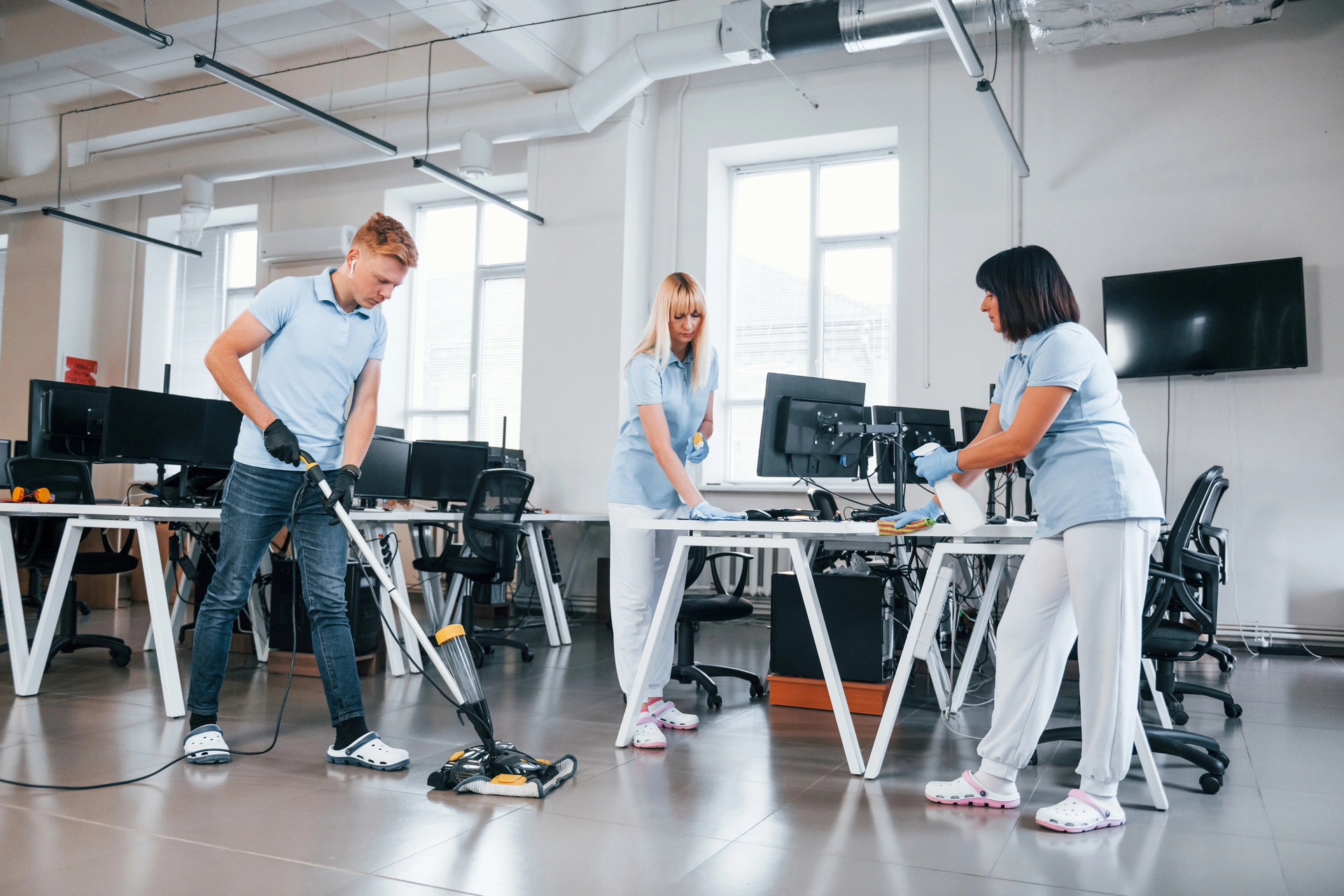 Three janitorial workers in uniforms are cleaning a modern office. One is vacuuming the floor, while the other two wipe surfaces on desks. The office is open-plan with large windows, computers, and modern furnishings.