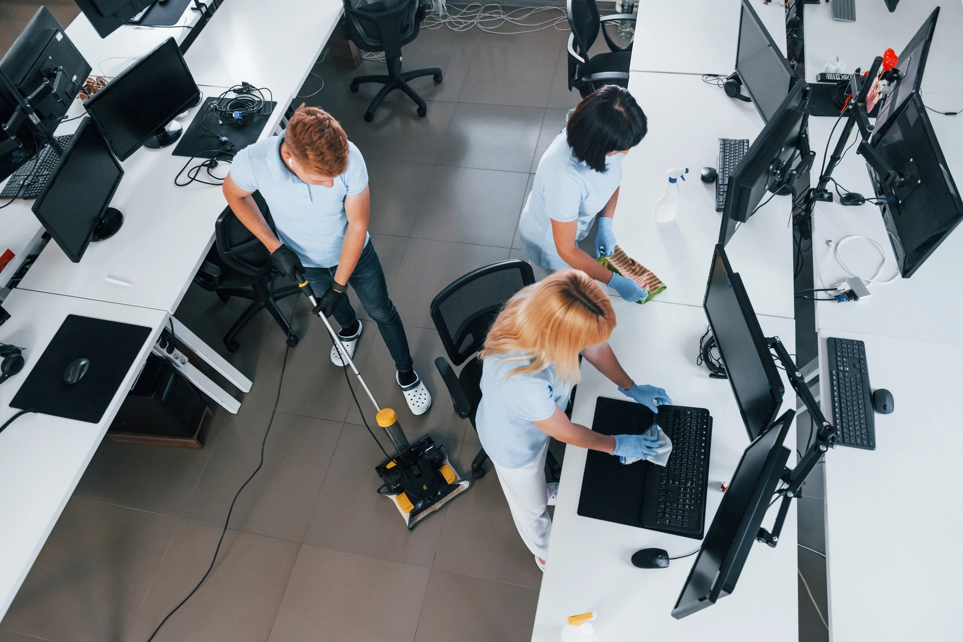 Three workers in blue uniforms clean an office. One mops the floor with a machine, while two others clean desks and computer screens with cloths. The office is bright and organized, with multiple desks and computers.